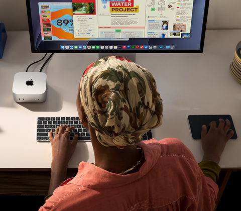 Una persona seduta a una scrivania che usa una Magic Keyboard e un Magic Trackpad per lavorare con un Mac mini collegato a uno Studio Display, accanto ci sono altri oggetti